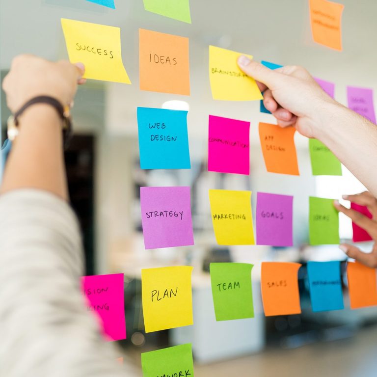 Brainstorming session with sticky notes on a glass wall. Text includes: SUCCESS, IDEAS, STRATEGY, GOALS, PLAN, and TEAM.